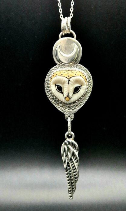 A silver pendant set with a small barn owl face hangs from a chain. It has a moon above the face, and a feather hanging from the bottom.