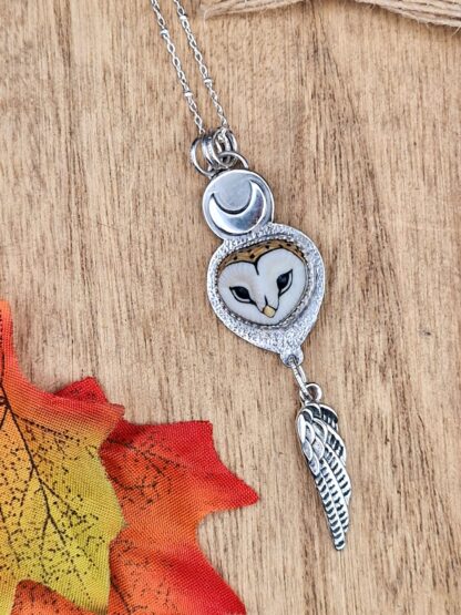 A silver pendant featuring a barn owl cabochon, a moon, and a feather set against a textured background.